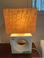 Lamp with square tan fabric lampshade with brown wavy lines, white ceramic lamp base with circular hole and size label