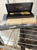 Photo showing Bombay pen set in black presentation box on top of three stacked books, on a metal shelf.