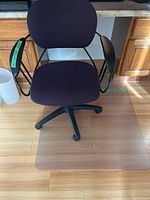 Office chair with black fabric and plastic armrests shown on clear floor mat on wood floor