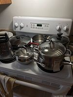 Image showing multiple pots, including two Lagostina pots with strainers and glass lids, two other pots, frying pans, and a box grater, all arranged on a white stove.