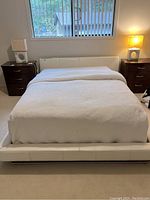 Front view of queen size white platform bed fully made with white coverlet, bedsides with lamps visible.