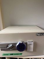 Front view of the compact white metal safe with digital keypad locking mechanism protected by a blue cover and handle. Shows some green tape with past code attempts written.