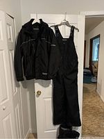 Full view of black Dakota ski jacket and bib pants hanging on door.