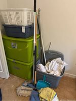 View showing stacked two large green storage bins, medium and smaller blue bins, white laundry basket on top, brooms, mop, curtain rods, and dusty mop and dustpan leaning against bins.