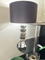 Full view of the lamp showing chrome pebble base and brown fabric shade on black shelf.
