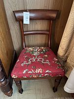 Antique wooden chair with red floral upholstered seat and curved backrest, placed against a wooden wall and next to a curtain.