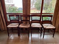 Four antique wooden chairs with woven wicker seats arranged side by side in front of a window.