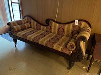 Side angle view of antique upholstered day bed showing carved wooden frame and patterned mattress with bolster pillows