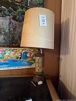 Full view of antique table lamp showing cylindrical base with floral art deco motif and beige fabric lampshade.