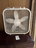 White square box fan on wooden table near curtain, showing front grill and blades