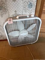 Photo of white box fan standing on wood floor with wallpaper background.