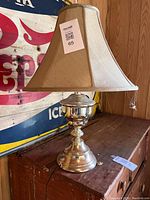 Photo showing the brass table lamp with beige fabric shade placed on a wooden surface next to a vintage wall sign. The lamp has a polished brass base and a classic shaped fabric shade.