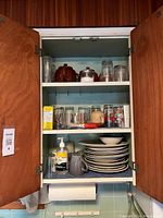 Open wooden kitchen cabinet showing two shelves with assorted kitchenware including plates, bowls, glasses, mugs, and containers.