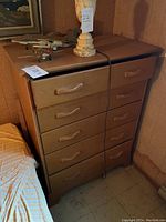 Wooden dresser with eight drawers seen from the front, placed in a room corner beside a bed. Top surface holds a lamp base and several small items.