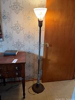 Full view of the 59-inch floor lamp showing the dark metal base, pole, and white decorative shade with the lamp turned on near a wooden door and wallpapered wall.