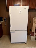 Closed white Kenmore refrigerator with freezer drawer below, showing exterior front and side sides against wooden paneled wall.