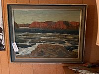 Framed landscape painting featuring a waterbody, rocky foreground, and reddish cliffs in the background