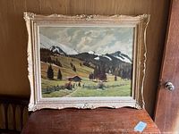 Framed landscape painting showing a cabin, pine trees, green fields, and snowy mountain peaks under cloudy sky.