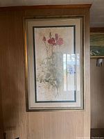 Framed large floral print on a wall, showing a vertical rectangular artwork with red flowers and green foliage in an abstract style. The frame is gold and ornate with a beige mat border.