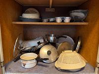 Shelf with various pots, lids, aluminum muffin tins, and a white plastic colander