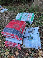 Several sealed bags of different soil types, including triple mix, black earth top soil, and possibly peat moss, lying on dry leaves outdoors.