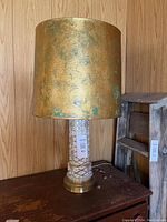Photo shows a white porcelain lamp base with gold lattice pattern and a gold metallic lampshade with patina marks on a wooden surface.