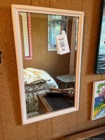 Photo of rectangular mirror with white frame hanging on a wooden wall, reflecting part of a room with furniture and wall art.