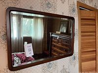 Wide rectangular wooden framed mirror hanging on a wall reflecting room interior including window with curtain and a dresser.