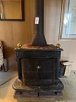 Front view of a black solid cast iron wood burning stove with brass finials and a glass door, attached to a tall chimney pipe.