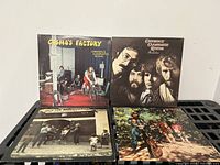Four vinyl LP albums by Credence Clearwater Revival showing front covers: Cosmo's Factory, Bayou Country, Willy and the Poor Boys, and Pendulum, placed on black plastic shelf.