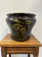 Front view of the dark glazed planter showing green leaf decoration and bulbous shape on wooden stool.
