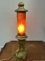 Front angle of the antique gothic table lamp lit, showing the warm orange mica shade and ornate base with gothic devil and gargoyle motifs.