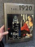 Front cover of the hardcover art book showing its title and artwork depicting city buildings and a woman in 1920s style clothing.