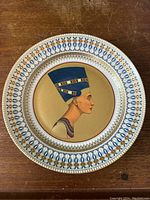 Top-down photo showing round porcelain wall charger with golden background and detailed profile of Queen Nefertiti in blue crown with decorative Egyptian style border in blue, gold and red.