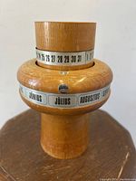 Close-up of wooden perpetual calendar showing the days of the month numbered 1 to 31 on the rotating metal band, along with months labeled in Hungarian (JÚNIUS, JÚLIUS, AUGUSZTUS, SZEPTEMBER) on another band around the rounded middle section.