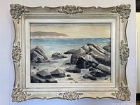 Frontal view of oil painting showing rugged coastal scene with rocks, ocean, distant hills, and signature by BV Paul. Surrounded by ornate wood frame with carved decorative elements.
