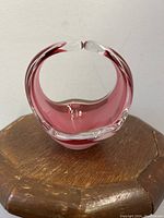 Opalescent cranberry art glass bowl with clear rim, showing side profile and sculptural curved design, placed on wooden surface.