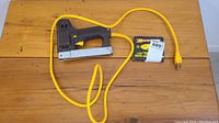 Electric staple tool with yellow cord on wooden surface, showing overall condition and included staple box.