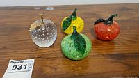 Four handmade glass apples on wooden surface, showing clear, red, green, and yellow apples with stems and leaves