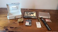 Seven assorted office items arranged on wooden surface: white metal cash box open, Seiko world clock with world map, clear acrylic display block, two letter openers, small gold-toned trinket box, compact calculator.