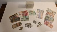 Overview photo showing assorted old banknotes and coins laid out with a wooden box in the background. Various international currencies visible, some notes folded and worn.