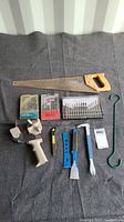Photo of assorted tools laid out showing saw, tape roller dispenser, drill bits packs, blue handled chisels, utility knife, and hook tool.