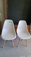 Two white modern dining chairs with molded plastic seats and wooden legs with metal rod supports, standing on a concrete floor with a dark background.