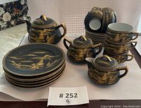 Full set of 23pc tea set showing teapot, creamer, sugar bowl, cups, saucers, and dessert plates arranged on white background with hand painted black and gold design.