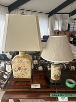 Two lamps on a wooden table in a living room setting; one lamp has a rectangular base with floral and bird designs, the other lamp has a cylindrical shape with floral and bird patterns. Cream-colored fabric shades on both.