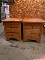 Front view of two oak nightstands side by side on concrete floor with particle board wall behind.