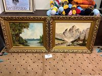 Two framed paintings side by side on the floor, one of a river landscape and other mountainous terrain.