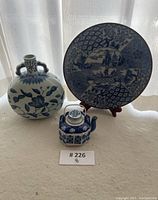 Set shows small hexagonal blue and white porcelain teapot with metal handle, round floral patterned vase, and decorative plate on wooden stand, all with blue and white Asian motifs.