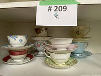 Multiple assorted fine and bone china teacups with saucers, showing various floral patterns and gold trimming, arranged in a shelf.