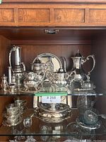 Full view of silver plated pitchers, coffee pots, sugar bowl, creamer, salt and pepper shakers, glassware and tray on glass shelves in china cabinet